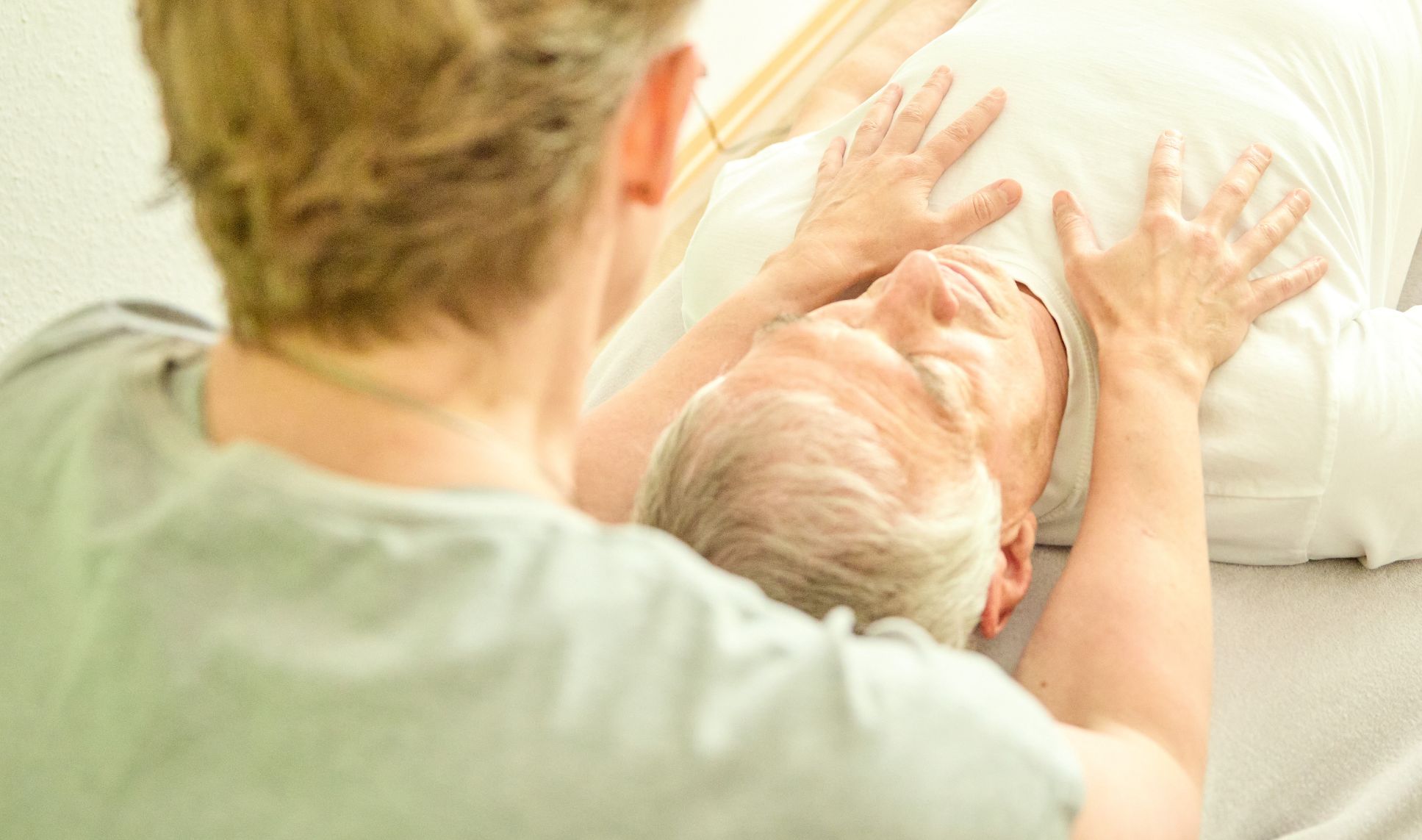 Älterer Herr, auf dem Rücken liegen, nur Oberkörper sichtbar, Hände der Osteopathin darauf
